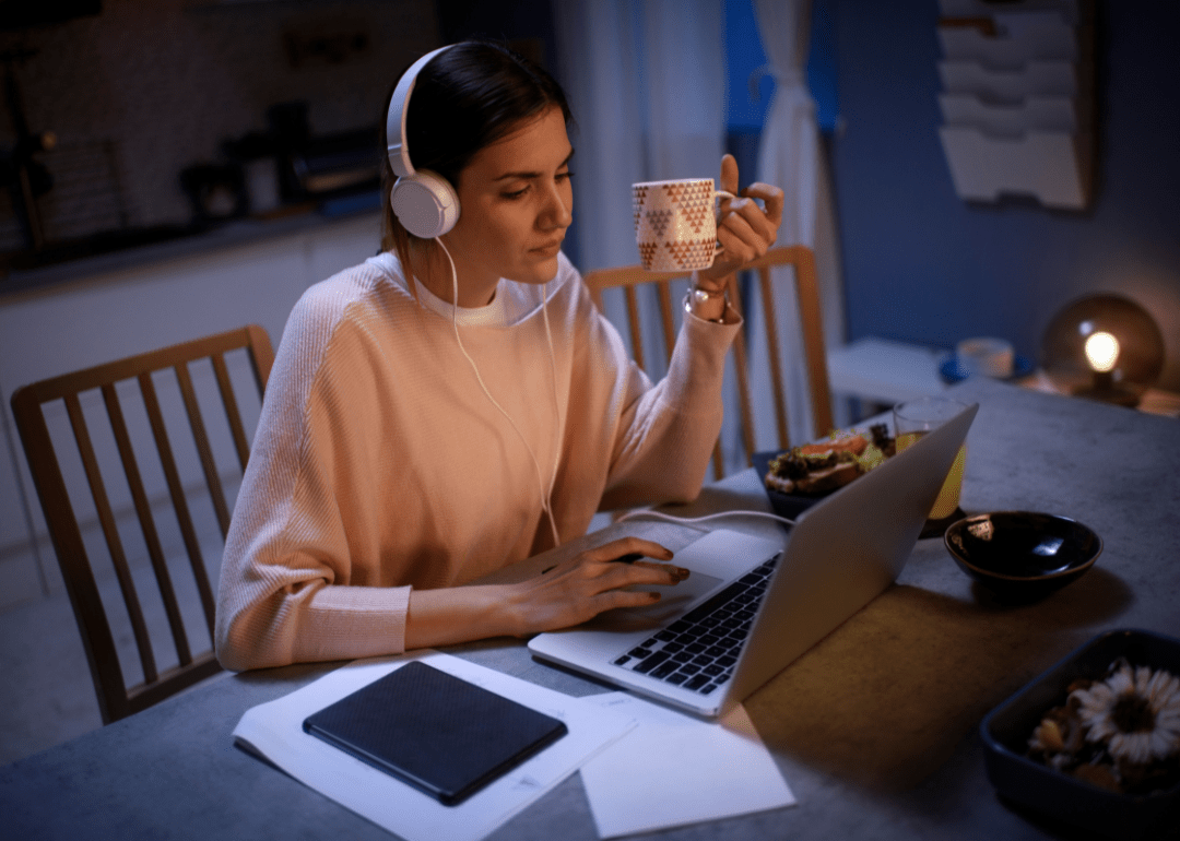 A woman with headphones on, holding a mug, and working on a laptop