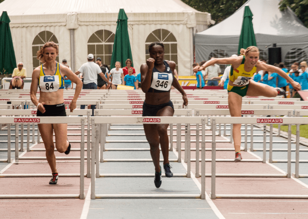 Three female athletes competing in a race and jumping over hurdles 