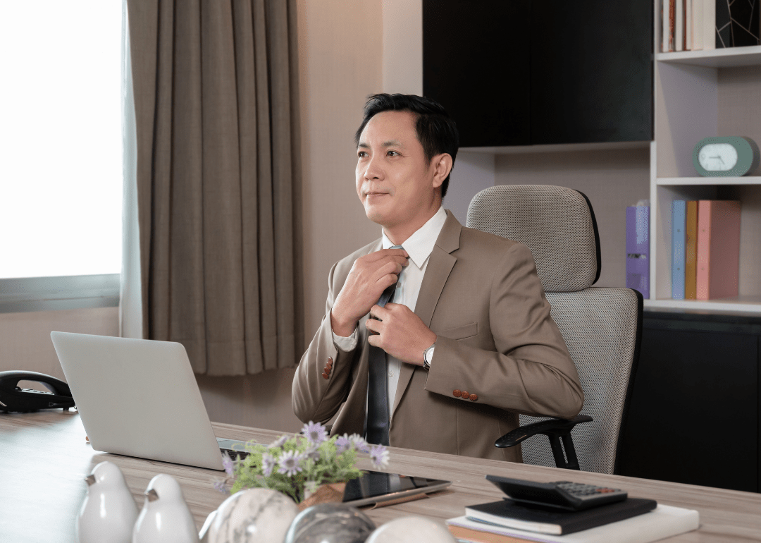 A man fixing his tie and sitting in a chair with a laptop on the desk